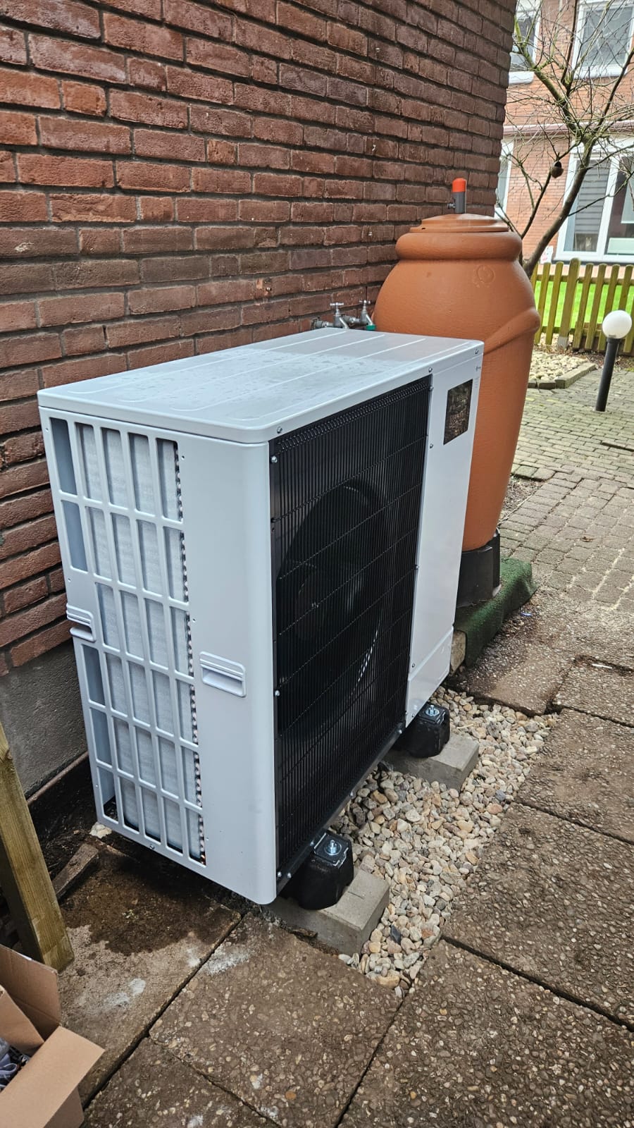 Weiße Wärmepumpe auf Kiesbett neben Backsteinwand, dahinter eine große Regentonne und Wasseranschluss.